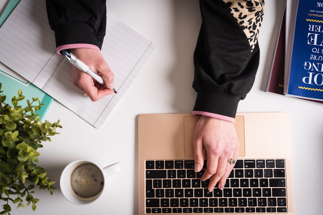 Adult writing symptom notes in a notebook beside a laptop and coffee